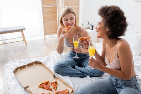 mujer rubia feliz comiendo deliciosa pizza cerca de amigo afroamericano con cóctel en copa de champán