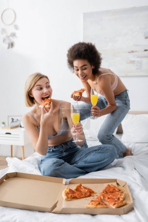 freudige afrikanisch-amerikanische Frau hält Cocktail in der Nähe einer blonden Freundin, die zu Hause im Bett Pizza isst