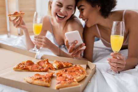 emocionadas mujeres multiétnicas con cócteles riendo cerca de teléfono inteligente y sabrosa pizza en el dormitorio