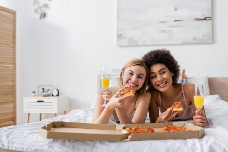 joyful multiethnic women looking at camera while lying on bed with delicious pizza and champagne glasses with cocktails