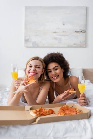 fröhliche interrassische Freunde mit leckerer Pizza und Cocktails in Sektgläsern auf dem Bett liegend und in die Kamera blickend