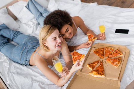 vista de ángulo alto de mujeres multiétnicas emocionadas sosteniendo deliciosa pizza y cócteles mientras yacen en la cama en casa