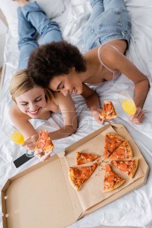 Blick aus der Vogelperspektive auf interrassische Frauen, die mit geschlossenen Augen in der Nähe von leckeren Pizza und Cocktails in Champagnergläsern lächeln, während sie im Bett liegen
