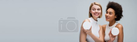 Téléchargez les photos : Pretty african american woman looking at blonde model posing with container of body cream isolated on grey, banner - en image libre de droit