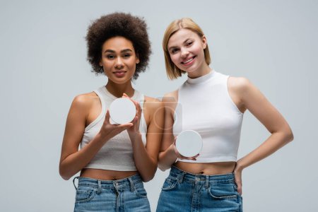 young interracial women in white tank tops holding containers with body cream and looking at camera isolated on grey