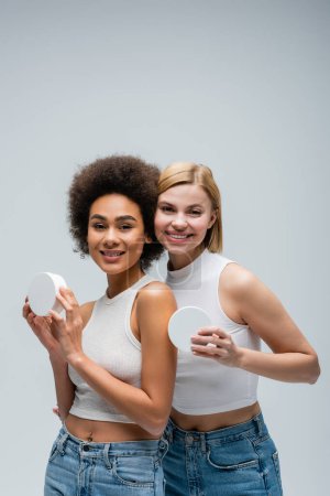 jolis modèles multiethniques en débardeurs blancs posant avec des récipients de crème cosmétique isolés sur gris