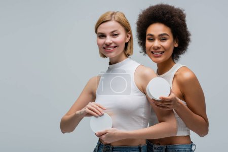 Téléchargez les photos : Blonde and brunette interracial women holding containers with cosmetic cream and smiling at camera isolated on grey - en image libre de droit