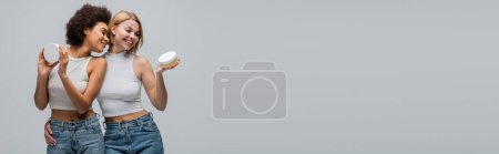 Téléchargez les photos : Modèle blond joyeux embrassant femme afro-américaine tout en tenant la crème cosmétique isolée sur gris, bannière - en image libre de droit