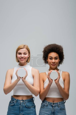 front view of young interracial women wearing white tank tops and holding jars with cosmetic cream isolated on grey