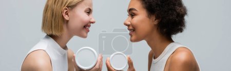 Téléchargez les photos : Side view of interracial blonde and brunette women with body cream smiling at each other isolated on grey, banner - en image libre de droit