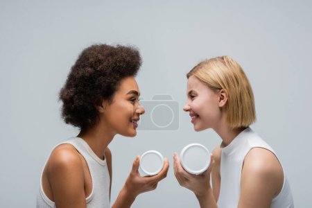 Téléchargez les photos : Side view of multiethnic blonde and brunette women holding cosmetic cream and smiling at each other isolated on grey - en image libre de droit