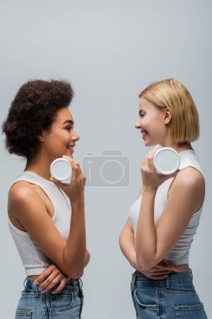Téléchargez les photos : Vue latérale de heureux modèles multiethniques exploitations conteneurs avec crème cosmétique isolé sur gris - en image libre de droit