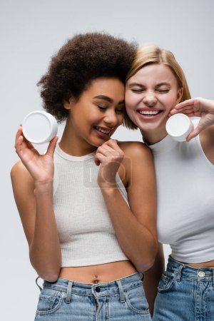 Téléchargez les photos : Jolies femmes interracial en hauts blancs posant avec de la crème cosmétique tout en souriant les yeux fermés isolés sur gris - en image libre de droit