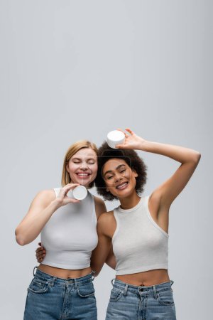 Téléchargez les photos : African american model hugging blonde woman posing with jar of cosmetic cream isolated on grey - en image libre de droit