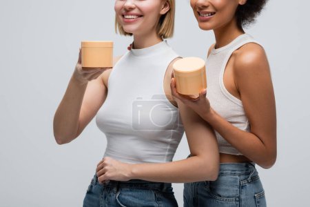 Téléchargez les photos : Cropped view of smiling interracial women in white tank tops posing with body cream isolated on grey - en image libre de droit