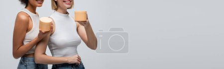 Téléchargez les photos : Partial view of young multiethnic models in white tops holding jars with cosmetic cream isolated on grey, banner - en image libre de droit