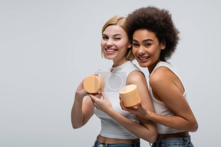 Téléchargez les photos : Cheerful interracial models in white tank tops smiling at camera while holding cosmetic cream isolated on grey - en image libre de droit