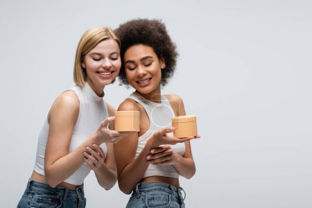 Téléchargez les photos : Pretty interracial women in white tops holding cosmetic cream and smiling isolated on grey - en image libre de droit