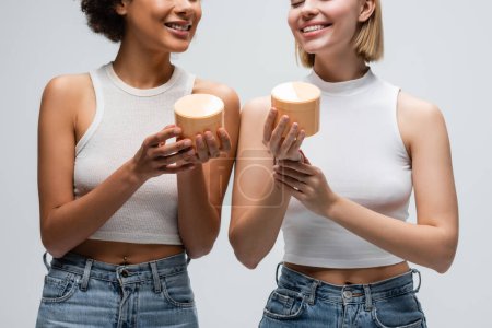 Téléchargez les photos : Vue recadrée de modèles interracial heureux en débardeurs blancs et jeans tenant crème pour le corps isolé sur gris - en image libre de droit