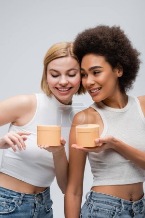 Téléchargez les photos : Jeune femme afro-américaine tenant des bocaux avec crème cosmétique près du modèle blond pointant du doigt isolé sur gris - en image libre de droit