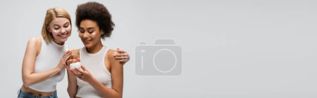 Photo for Smiling blonde model touching shoulders of african american woman holding face cream isolated on grey, banner - Royalty Free Image
