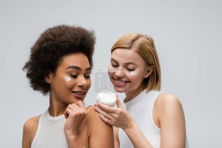 Téléchargez les photos : Heureux femmes multiethniques en hauts blancs regardant récipient avec crème visage isolé sur gris - en image libre de droit