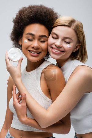 Photo for Sensual blonde model holding face cream near smiling african american woman isolated on grey - Royalty Free Image