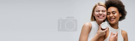 Téléchargez les photos : Joyeux modèles interracial souriant avec les yeux fermés près du bocal avec crème visage isolé sur gris, bannière - en image libre de droit