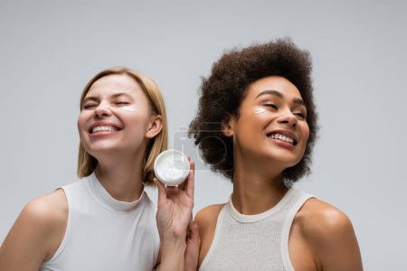 modelos interracial complacidos en tapas blancas posando cerca de tarro de crema facial aislado en gris