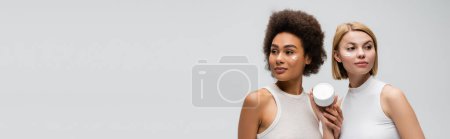interracial blonde and brunette women looking away near jar of face cream isolated on grey, banner