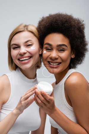 Photo for Joyful interracial women in white tank tops looking at camera near jar of face cream isolated on grey - Royalty Free Image