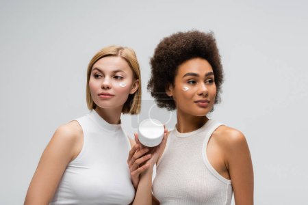 mujeres jóvenes interracial en camisetas blancas mirando hacia otro lado cerca del frasco con crema cosmética aislada en gris