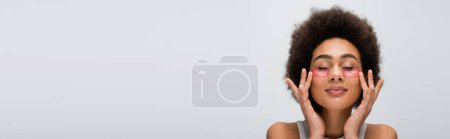 Foto de Morena mujer afroamericana con los ojos cerrados aplicando parches levadizos aislados en gris, pancarta - Imagen libre de derechos