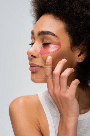 Foto de Young african american model with closed eyes and collagen eye patches holding hand near face isolated on grey - Imagen libre de derechos