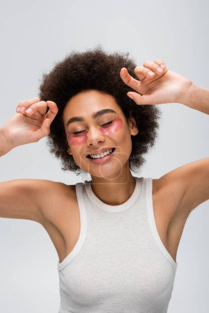 joyful african american woman in white tank top and collagen eye patches posing with closed eyes isolated on grey