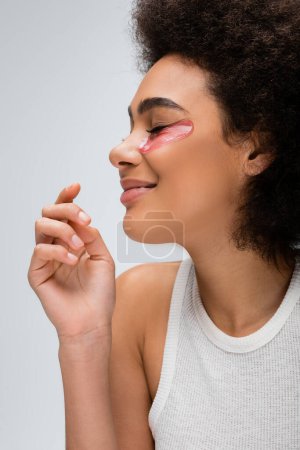 profile of african american woman with collagen eye patch smiling with closed eyes isolated on grey