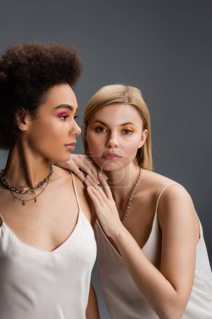 blonde model with colorful makeup looking at camera near young african american woman in silver necklaces isolated on grey