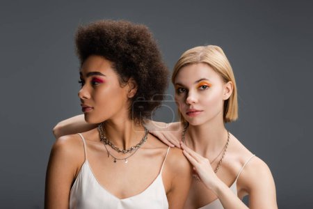 pretty blonde woman with colorful visage looking at camera near african american model looking away isolated on grey