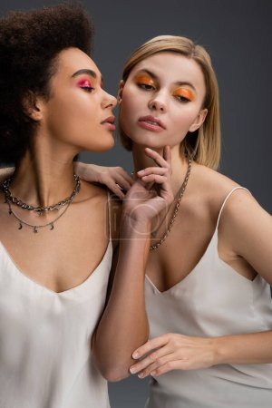 sensual african american woman in white strap dress and silver necklaces touching chin of blonde model with bright makeup isolated on grey
