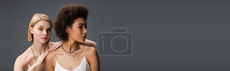 young african american model in white strap dress looking away near blonde woman with bright makeup isolated on grey, banner