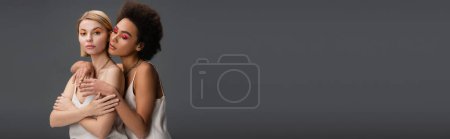 Téléchargez les photos : Young african american woman hugging blonde model in white strap dress and bright visage isolated on grey, banner - en image libre de droit