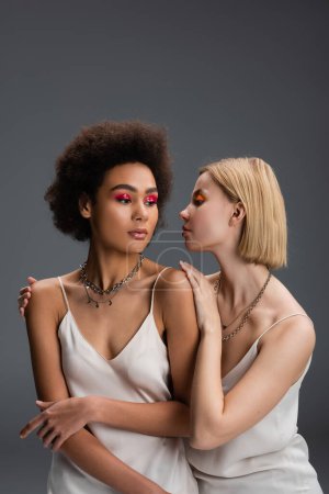 blonde woman touching shoulders of african american model in white strap dress and silver necklaces isolated on grey