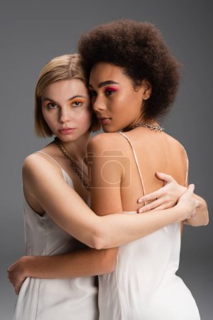 sensual interracial women in white strap dresses and colorful makeup embracing isolated on grey