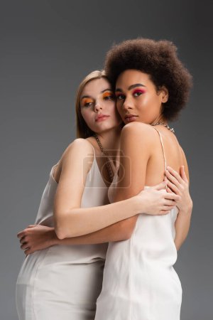 multiethnic blonde and brunette models in white camisoles and bright visage embracing isolated on grey