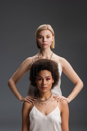 stylish interracial women in white strap dresses and colorful makeup looking at camera isolated on grey