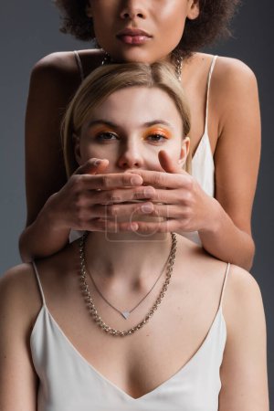 Foto de African american woman covering mouth of blonde model in white strap dress and silver necklaces isolated on grey - Imagen libre de derechos