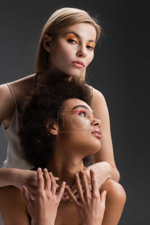 blonde model with orange eye shadows embracing brunette african american woman while looking at camera isolated on grey