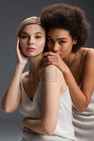 pretty multiethnic women with colorful visage posing in white strap dresses and looking at camera isolated on grey