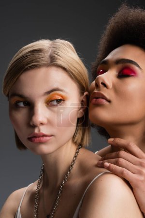 blonde woman with orange eye shadows looking away near sensual african american model posing with closed eyes isolated on grey