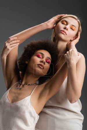 stylish and sensual interracial models in white camisoles and bright makeup posing with closed eyes isolated on grey